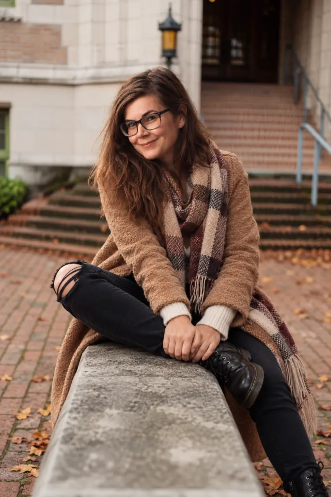 Junge Frau mit braunen Haaren und Brille sitzt auf einer Betonbank in einem Innenhof. Sie trägt einen beigen Cardigan, kariertem Schal, schwarze Jeans