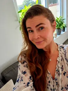 Frau mit langen rötlich-braunen Haaren und geblümtem Oberteil lächelt in die Kamera, im Hintergrund Pflanzen auf einer Fensterbank.
