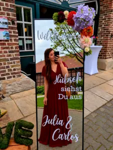 Hochzeitsdeko mit Spiegelbild einer Frau im roten Kleid, Willkommensschrift und bunten Blumenarrangements an Backsteinhaus.